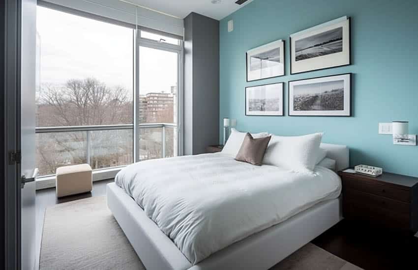 Bedroom with pale blue accent wall, bedding, pillows, window, nightstand, and ottoman